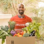 Deliveryman in red shirt holding a box of fresh vegetables, smiling at the doorway.