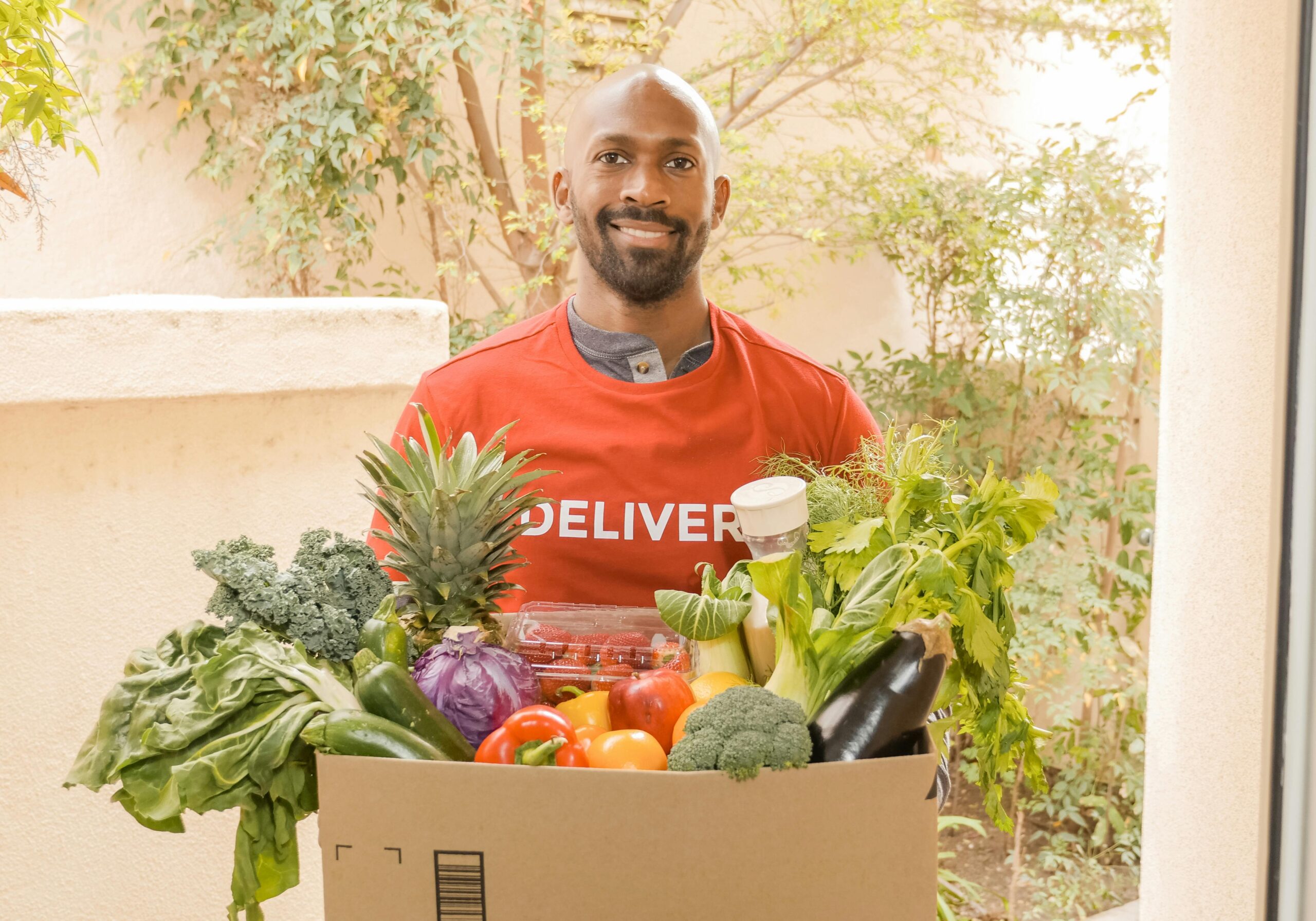 Deliveryman in red shirt holding a box of fresh vegetables, smiling at the doorway.